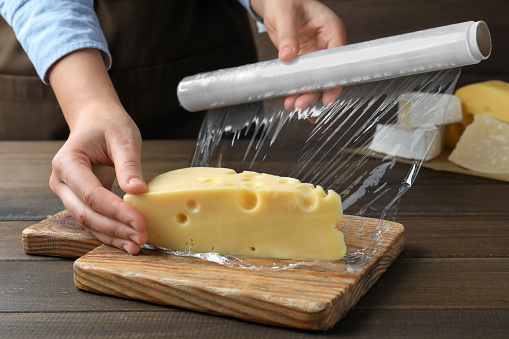 Woman putting plastic food wrap over block of cheese at wooden table, closeup