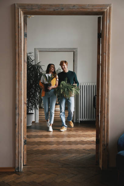 Happy young couple holding houseplants while walking by their new apartment together stock photo