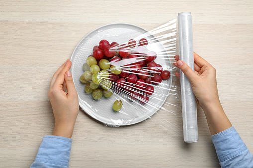 Woman putting plastic food wrap over plate of fresh grapes at wooden table, top view