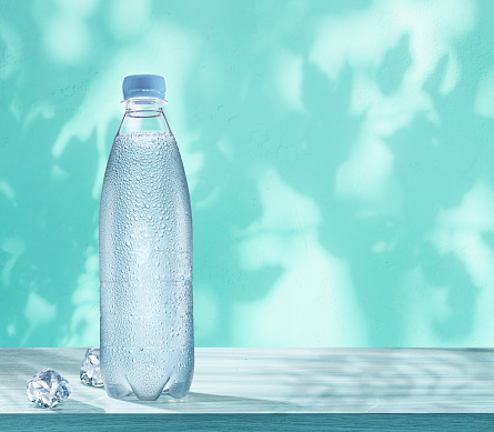 Plastic bottle of chilled water with condensation drops and two ice cubes on the table. Wall with leaf shadows at the background.