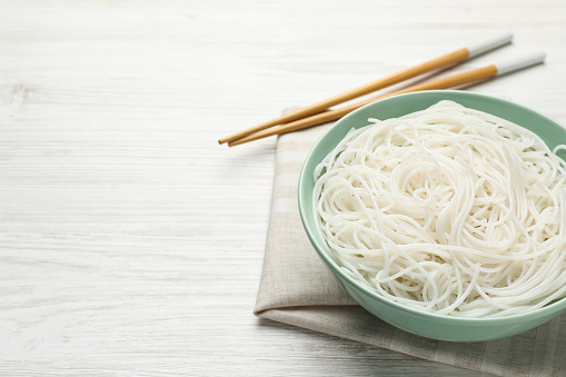 Bowl with cooked rice noodles and chopsticks on white wooden table. Space for text