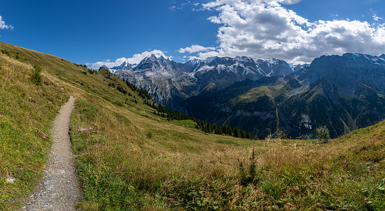 A woman with a hiking backpack hiking on a narrow pathway in high Italian Dolomites. Steep, sharp mountain chain in the back. She is crossing a lush green meadow. Discovering and exploring the nature