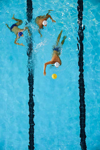 A forward water polo player drives the ball. A defender and a teammate fallow him.