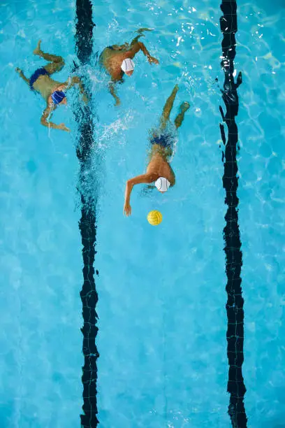 A forward water polo player drives the ball. A defender and a teammate fallow him.