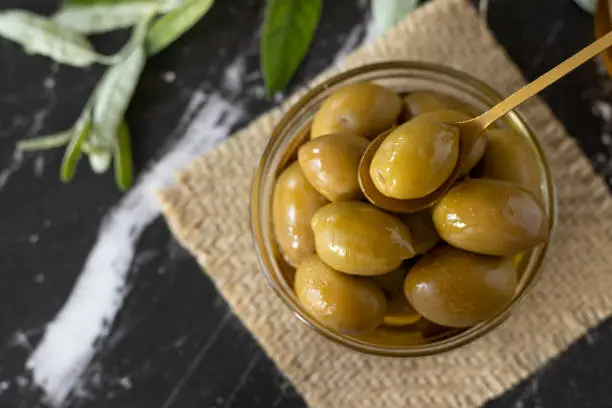 Fresh olives in a glass bowl with a golden spoon on a dark textured background with a green olive branch. Top table view. A closeup.