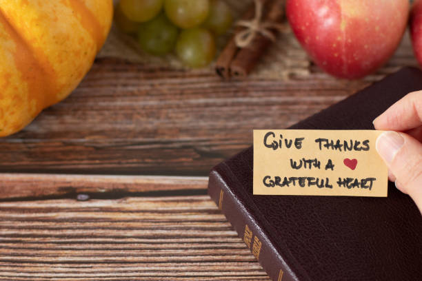 Hand holding a handwritten thanksgiving note to give thanks with a grateful heart with closed Holy Bible Book and fresh autumn fruit on wooden background with copy space stock photo
