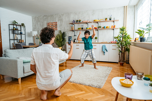 Young father filming his child dancing in the living room using smart phone.