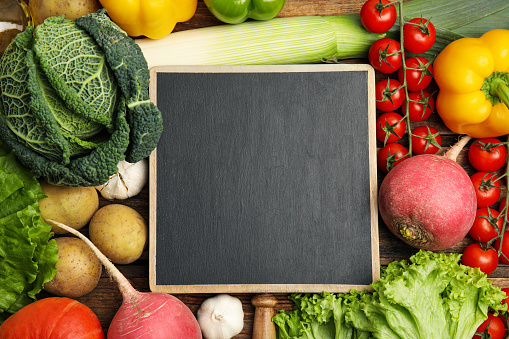 Blank chalkboard surrounded by different fresh vegetables on wooden table, flat lay with space for text. Cooking classes