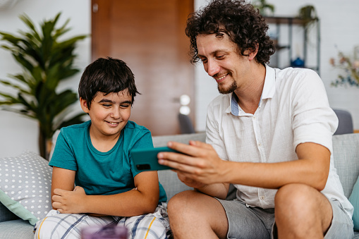 Young father sitting on the sofa in the living room with his young son and using a smart phone.