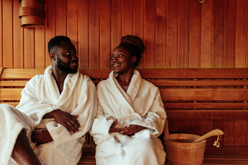 A happy African American couple is in the sauna. They are wearing bathrobes and are lovingly looking at each other.