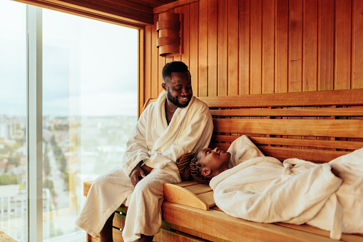A young African American couple are on their honeymoon. They are having a spa day in the resort. They are spending time in the sauna. The husband is seated and his wife is lying on the bench.