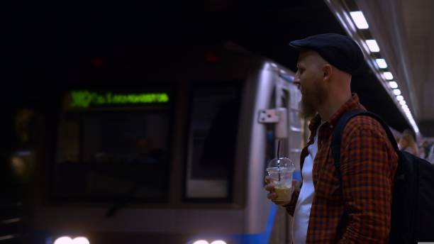 A bearded man in a cap stands on a subway platform and drinks iced coffee while waiting for a subway train. A typical day on the subway, a man goes to work every day early in the morning.
