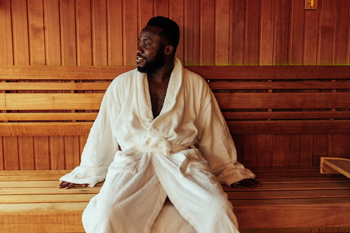 A young and happy African American man is sitting in his bathrobe in sauna enjoying his day.