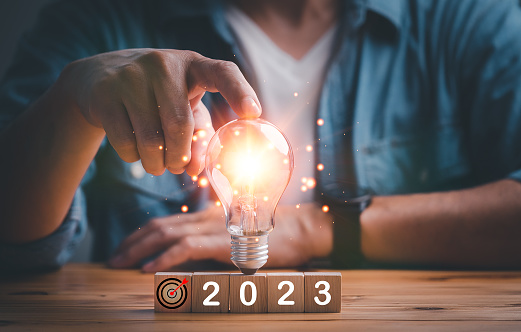 2023 New Year Goal Target, Action, Plan, Idea, Inspiration Concept. Businessman touching light bulb with grow light sparkling effect on wood cube with target symbol 2023 dark background. Concept 2023
