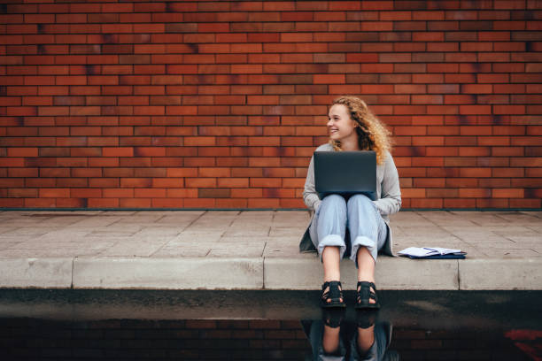 istock Young curly haired university student using laptop sitting on street near university campus on brick wall with copy space. Concept of e-learning, distance study or remote learning concept. Youth lifestyle. Education internet technology. Beauty portrait. 1428744286