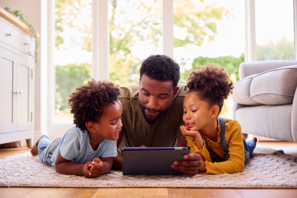 自宅で父親は、デジタルタブレットを使用して子供たちとラウンジで敷物の上に横たわっている - digital tablet looking child offspring ストックフォトと画像