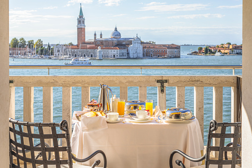 Venice, Italy - September 21, 2022: Breakfast in the room table setting is awaiting the guests on the balcony of one of the Hotel Danieli's room in Venice, Italy. In the background the lagoon and the San Giorgio Maggiore Island is visible.\nVenice is built on a group of 118 small islands separated by canals that and linked together by over 400 bridges.