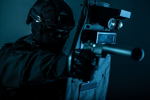 Police special forces fighter, assault team member, tactical group officer in black uniforms, aiming with silenced pistol while hiding behind, covering himself with ballistic shield, toned colorized