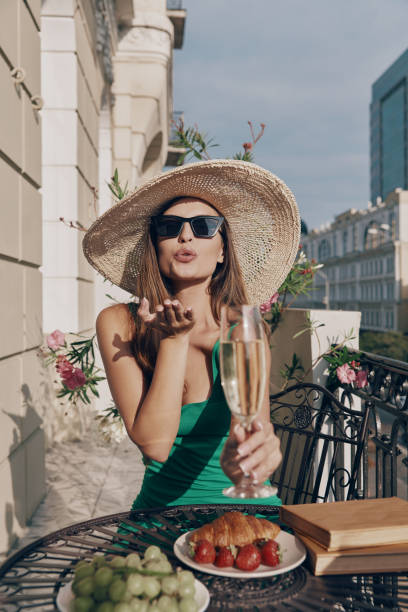 Beautiful young woman holding flute with champagne and blowing a kiss while relaxing on the balcony stock photo