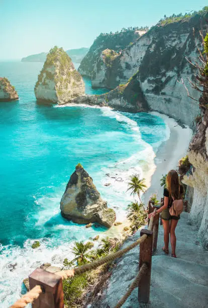 Photo of Travel girl with backpack in shorts on the ocean, cliffs and tropical beach background. Nusa Penida, Indonesia. Female silhouette, blonde hair.