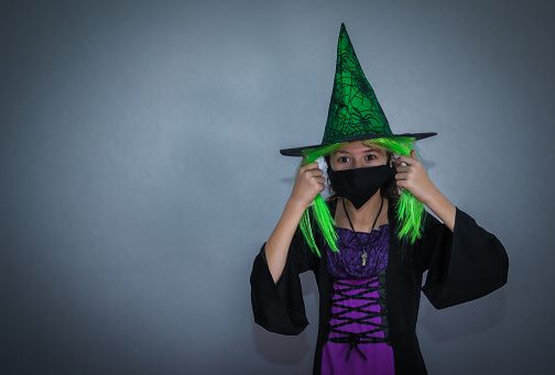Portrait of a smiling caucasian girl in dress, halloween hat with green hair and black mask, looking at the camera and standing on the right against the wall on a gray background with copy space on the left, close-up side view. Halloween concept, Halloween celebration.