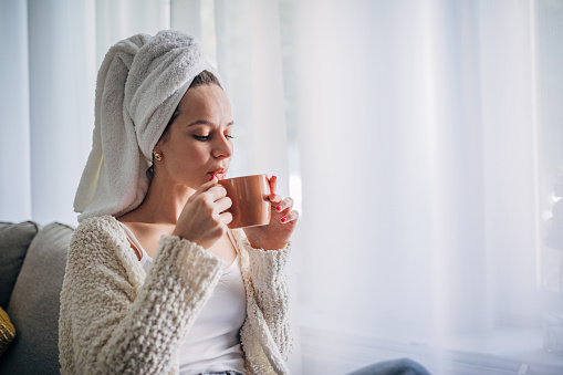 One woman, beautiful young woman sitting on sofa in living room and holding a cup of coffee, she has a towel on her head.