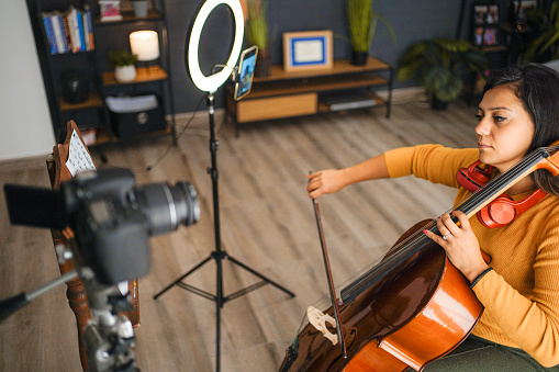 Young woman filming herself while playing violoncello at home