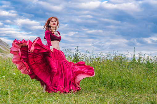Young beautiful and happy red-haired girl dances incendiary dance in red ball gown on areen meadow against blue cloudy summer sky.