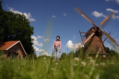 A black-haired woman stands in a green meadow on a clear summer day against the backdrop of a mill and sky, viewed from below from the grass. The girl is dressed in a pink shirt and blue jeans, her hands are down and she looks tired. Selective focus.