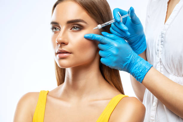 Cosmetologist makes rejuvenating anti wrinkle injections on the face of a beautiful woman in a yellow blouse. Female aesthetic cosmetology in a beauty salon. stock photo