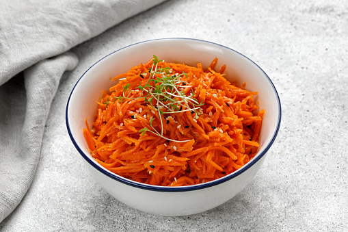 Korean Asian carrot salad with sauce and spices in a bowl. Vegetable salad with carrot, sesame seeds and microgreens. Grey background, copy space, text.