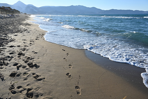 Sandy beach with footprints in the wet sand. Soft waves are washing away the footprints. There are some defocused beach umbrellas and incidental people on the background. Mountains are over horizon.