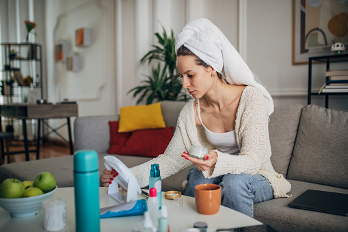 One woman, beautiful young woman sitting on sofa in living room and woman applying face cream on her face, she has a towel on her head.