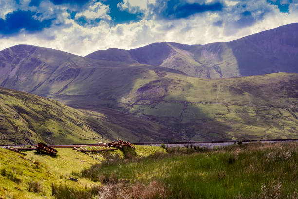 Mt Snowdon, Wales. Photo taken about half way up the climb. A rustic railway sweeping from the foreground to the mid ground. A rolling mountainside with lush greens and browns. A deep blue sky and cloudy sky casting shadows on the ground.