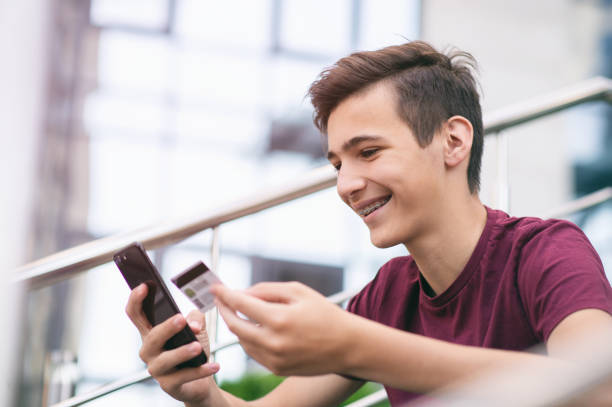 Teenage boy with a credit card and mobile phone makes purchasing outdoors. Happy young man is using smartphone and bank card for online shopping. Handsome smiling guy holds bank card and cell phone stock photo