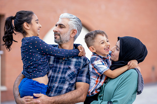 A small Middle Eastern family of four spend time bonding together in their new country after immigrating to Canada.  The mother and father are each holding a child in their arms, are each dresed casually and the mother has a Hijab on as they laugh and spend quality time together downtown.