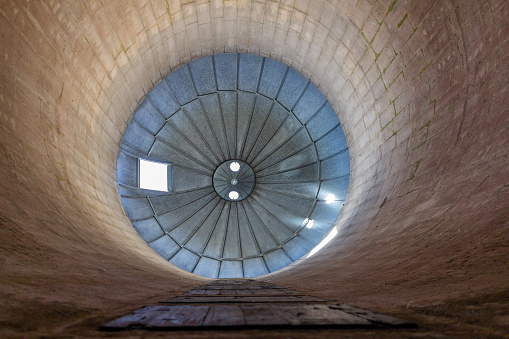 This image shows an abstract low-angle texture background view of the interior of a vintage round farm silo, with cement block walls and a dome shaped steel metal roof emitting ambient sunlight.