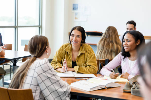 Female friends smile while listening to unrecognizable female friend Taking a break from their assignment, the two young adult female friends smile as they listen to the unrecognizable young adult woman seated across from them. female high school student stock pictures, royalty-free photos & images