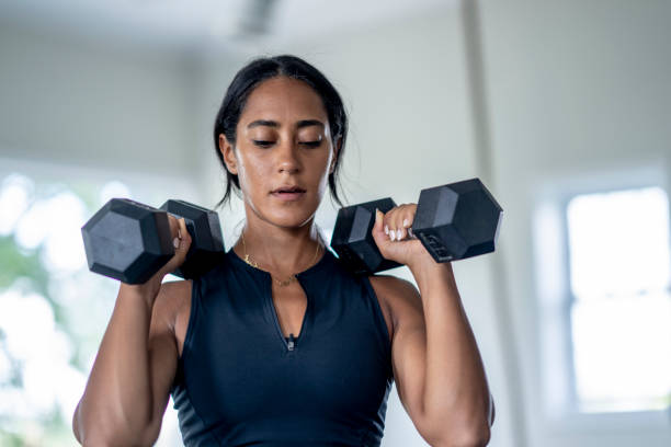 Woman Weight Lifting A young woman of Middle Eastern decent stands with a dumbbell in each hand as she lifts the weights during a workout. She is dressed comfortably in athletic wear and focused on her repetitions. home workout stock pictures, royalty-free photos & images