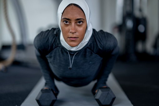 A young woman of Middle Eastern decent lays out on a yoga mat in a fitness center as she works out by herself.  She is dressed comfortably in athletic wear and is focused on lifting the weights in her hands as she holds a push up position.