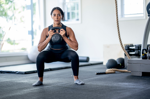 A young woman of Middle Eastern decent stands with her feet wide apart and a kettlebell in her hands as she works out alone at home.  She is dressed comfortably in athletic wear and focused on her breathing.