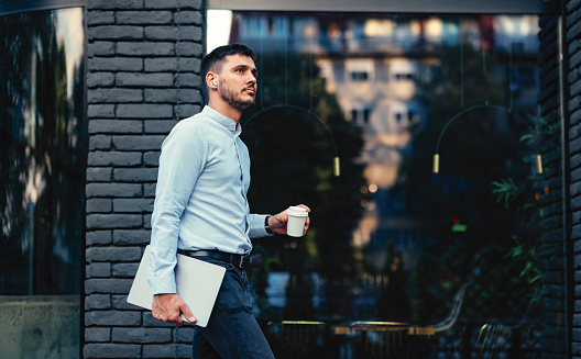 Handsome man holding laptop computer and disposable cup of coffee while walking outdoors.