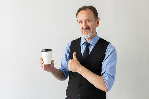 Portrait of satisfied mature businessman holding coffee cup and making thumb up gesture. Senior Caucasian manager wearing formalwear advertising takeout drink. Coffee break concept