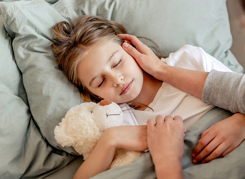 Cute child girl hugging teddy bear toy and sleeping in the bed. Mother hands cares about kid daughter during napping