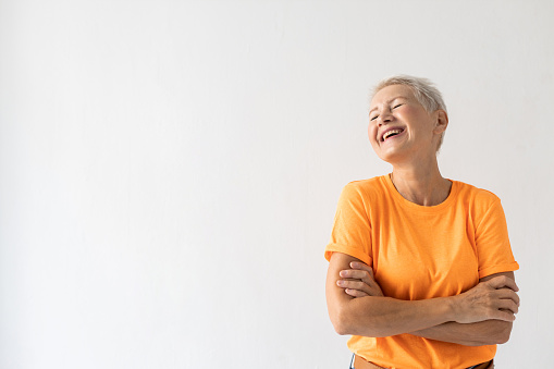 Portrait of senior woman laughing. Female model in orange T-shirt standing with hands crossed laughing hard. Portrait, studio shot, happiness concept