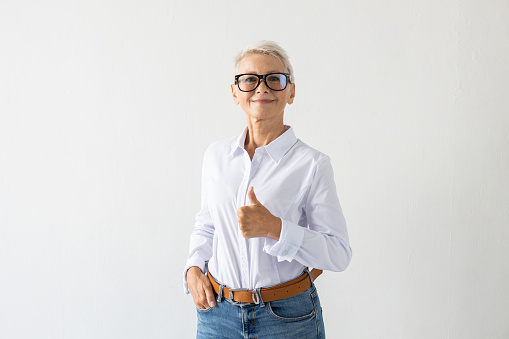 Portrait of senior woman with thumbs up. Female model in white shirt making thumbs up gesture, approving. Portrait, studio shot, approval concept