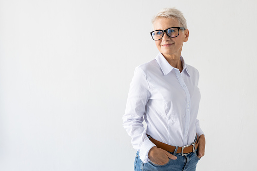 Portrait of senior woman standing proudly. Female model in white shirt standing holding hands in pockets. Portrait, studio shot, pride concept