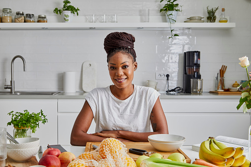 Portrait of a beautiful young adult black woman at the kitchen table, representing a healthy lifestyle, with a lot of fresh organic fruit and vegetables on it, for a healthy breakfast and juice preparation, vegan or vegetarian food