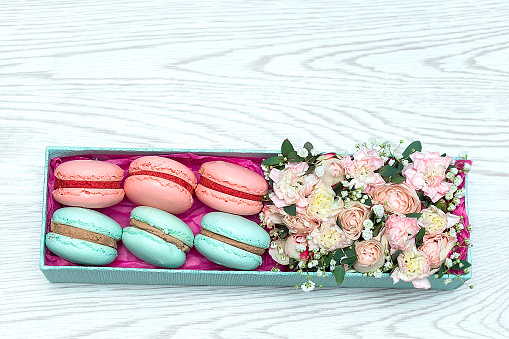 Pink and blue flowers and sweet macaroons in gift box on white wooden background with copy space for text. Top view flatlay. Roses and dessert as romantic present.
