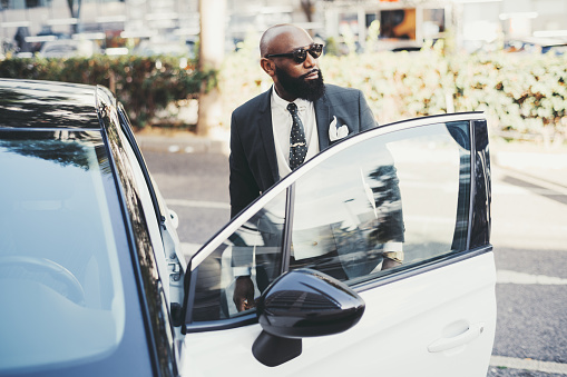 A portrait of a tough bald African businessman with a beautiful black beard, in sunglasses and a tailored fashionable costume, looking aside while entering his modern glossy car parked on the street